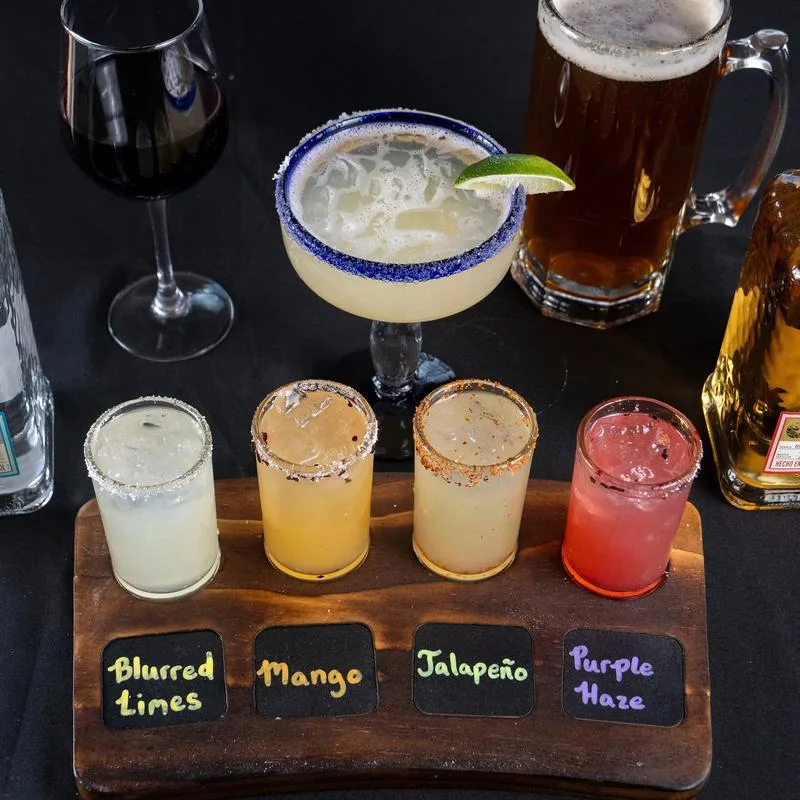 Four colorful margaritas in a tasting flight with a larger margarita, beer, and wine behind them.
