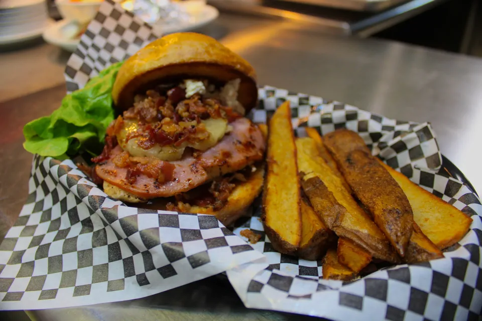 A stacked burger with bacon, lettuce, and pineapple served beside potato wedges.