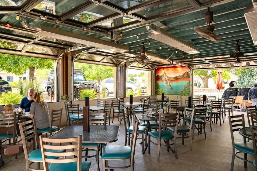 Open-air dining room at Coyote’s with raised glass garage doors, teal chairs, and a colorful mural.