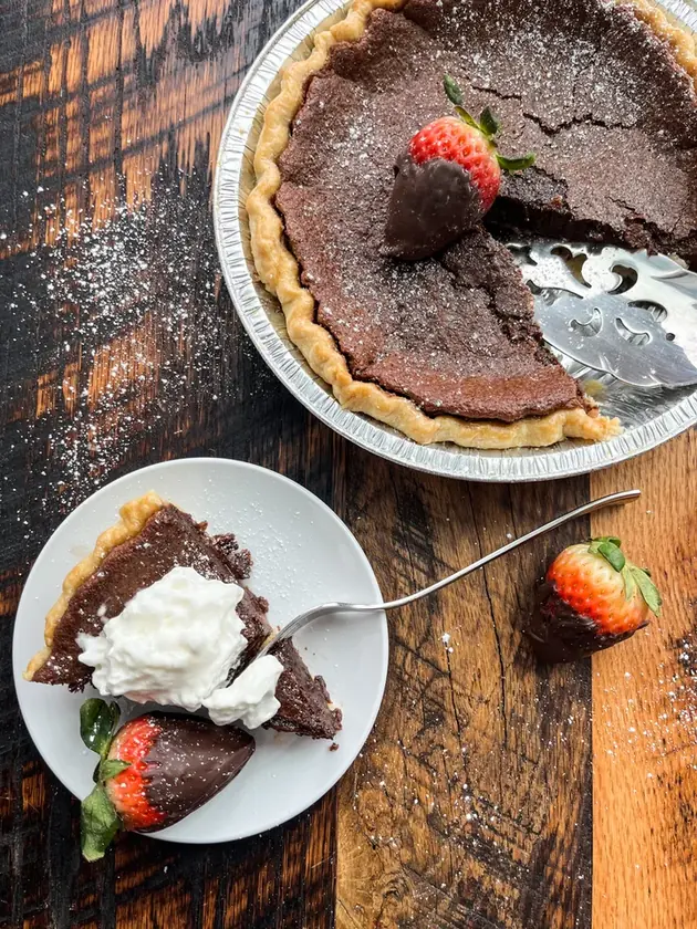 Chocolate pie and a plated slice with whipped cream and chocolate-dipped strawberries on a wooden table.