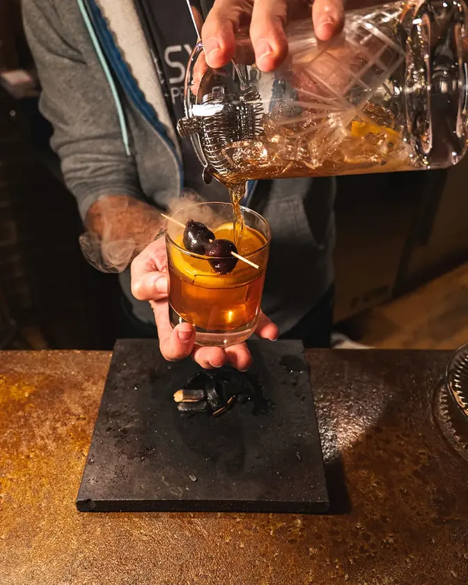 A bartender pouring a smoked amber cocktail over an orange slice and cherries at Syntax.