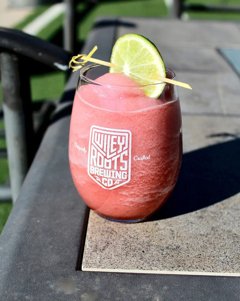 A pink frozen drink with a lime garnish in a Wiley Roots Brewing glass on a patio table.